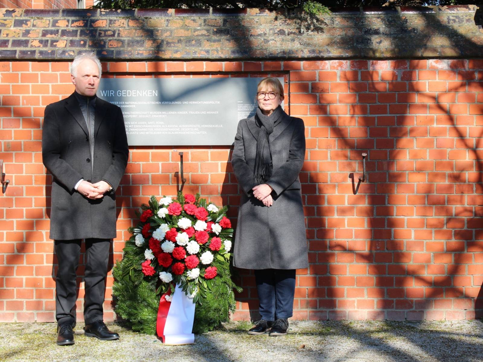 Eine Frau und ein Mann stehen vor einer Gedenktafel. Sie sind in Schwarz gekleidet und zwischen ihnen ist ein Kranz.